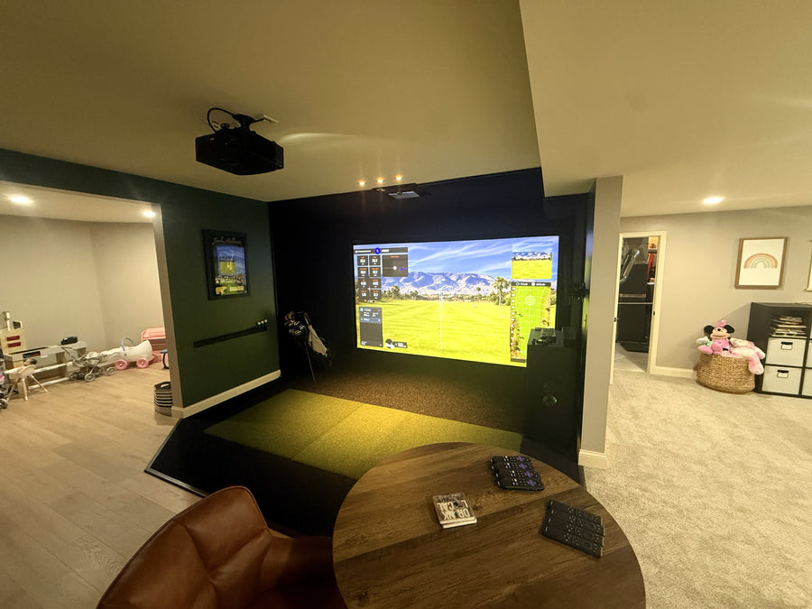 Indoor golf sim room with a large screen displaying a golf game, surrounded by furniture and decor.