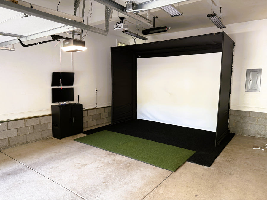 Garage golf simulator setup with screen and green mat on a concrete floor.