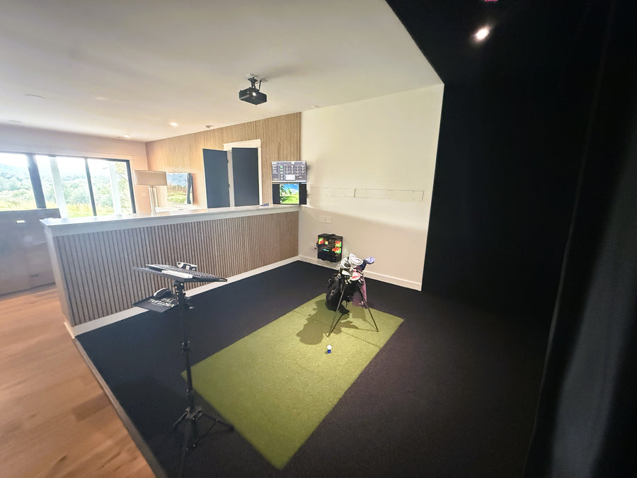 Indoor golf simulator setup in a room with a green turf area and various equipment.
