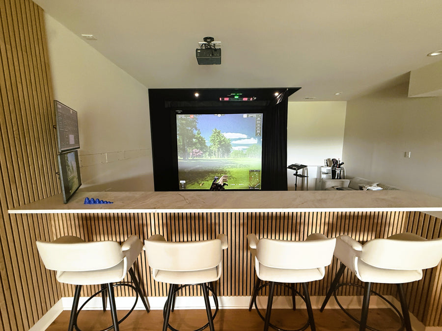 Golf simulator room with a large screen displaying a video game, surrounded by white chairs.