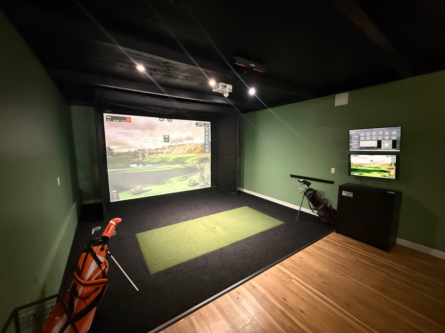 Golf simulator room with screen displaying round of golf, golf club on the side and table and seating in the back.