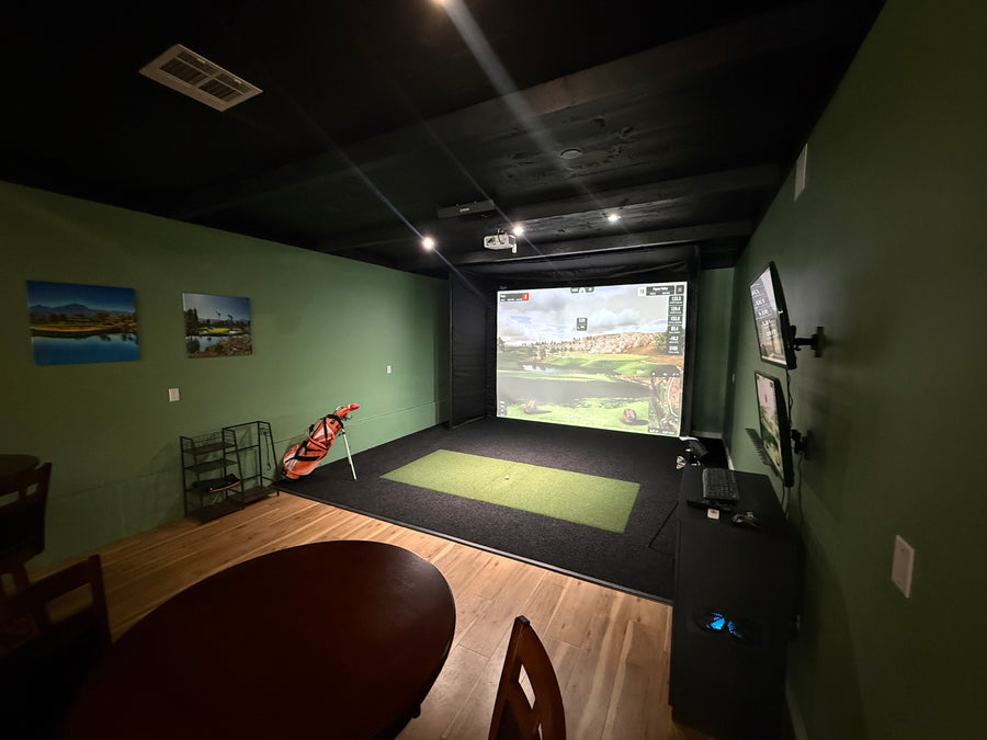 Golf simulator room with screen displaying round of golf, golf club on the side and table and seating in the back.