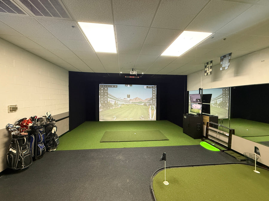 Room with golf simulator on back wall displaying golf course round. Golf clubs on the side and putting green on the floor in the back.
