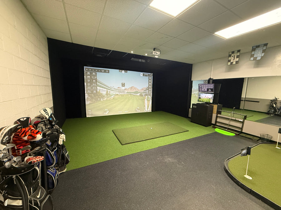 Room with golf simulator on back wall displaying golf course round. Golf clubs on the side and putting green on the floor in the back.