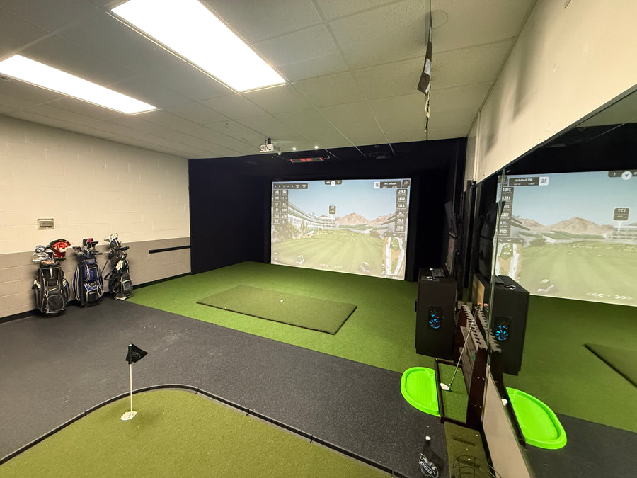 Room with golf simulator on back wall displaying golf course round. Golf clubs on the side and putting green on the floor in the back.