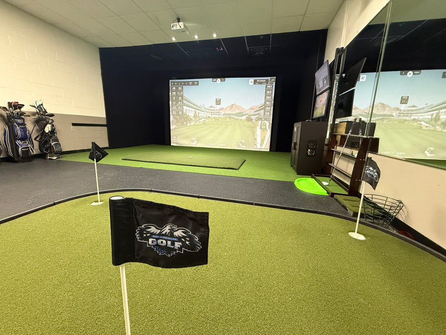 Room with golf simulator on back wall displaying golf course round. Golf clubs on the side and putting green on the floor in the back.