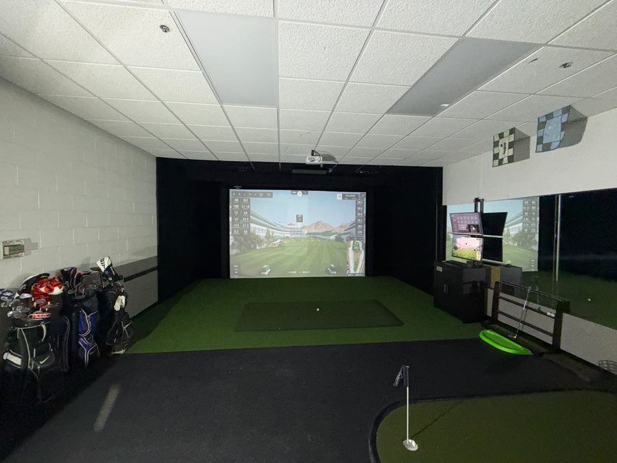 Room with golf simulator on back wall displaying golf course round. Golf clubs on the side and putting green on the floor in the back.