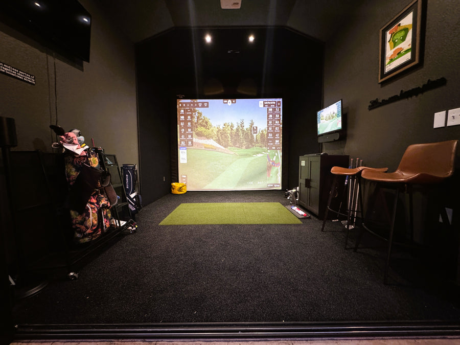 Indoor golf simulator setup with screen displaying a golf course, surrounded by chairs and equipment.