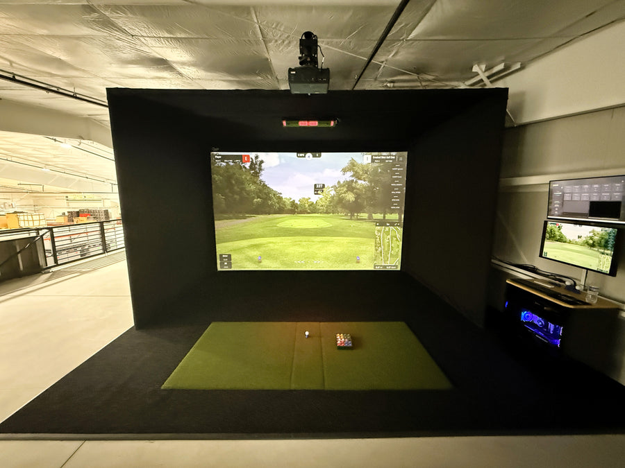 Golf simulator setup with screen displaying a golf course and various equipment on the floor.