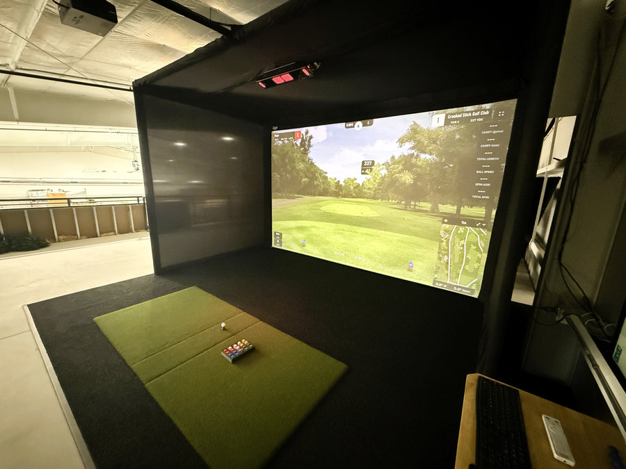 Golf simulator setup with screen displaying a golf course and a mat on the floor.