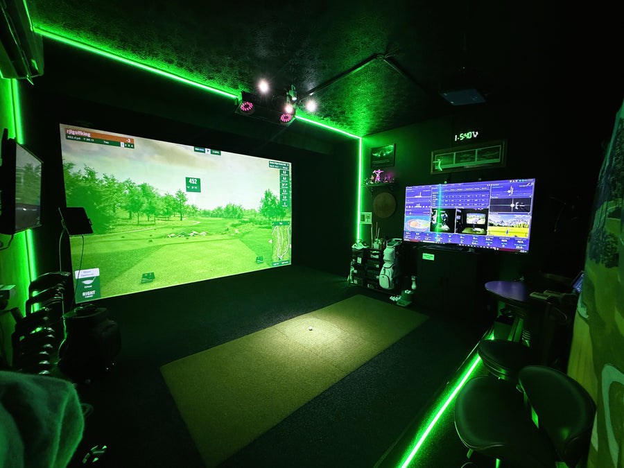 A dark indoor golf simulator room with a large projection screen showing a golf course, green LED neon lighting along the ceiling and floor, a driving mat, and multiple monitors and seating areas.