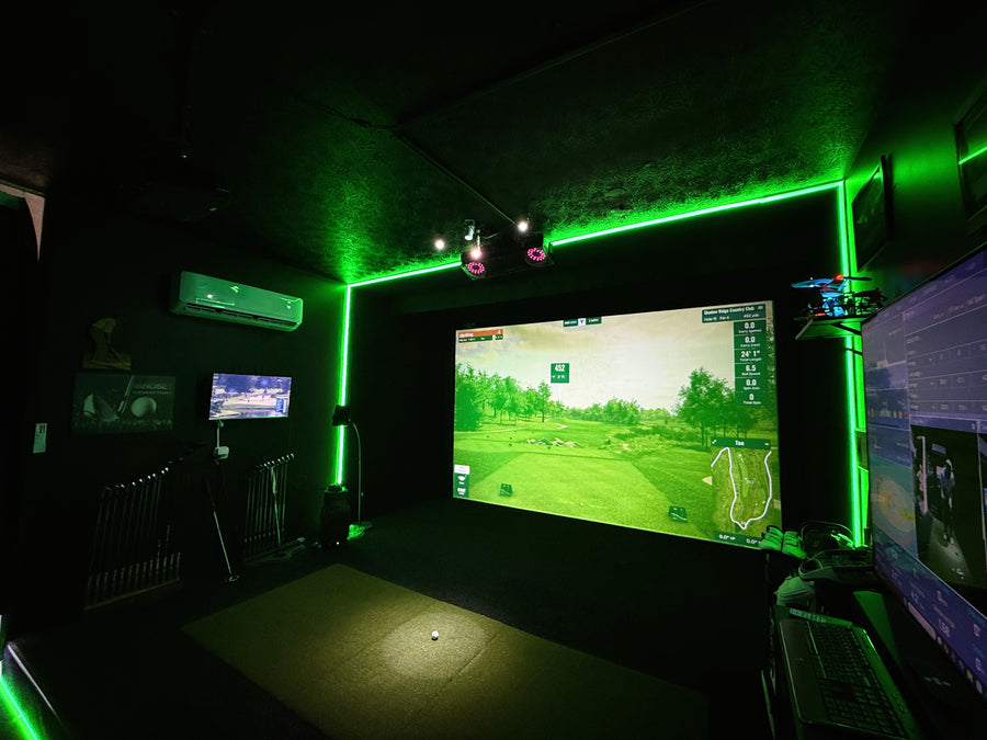 A dark indoor golf simulator room with a large projection screen showing a golf course, green LED neon lighting along the ceiling and walls, a hitting mat on the floor, and multiple monitors on the right side.