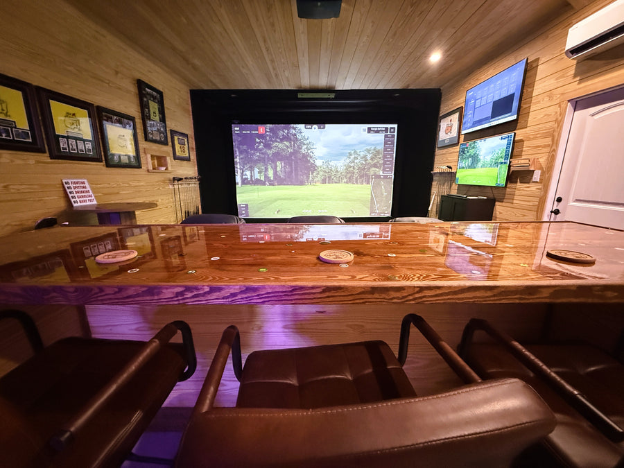 Indoor golf simulator area with a large screen displaying a sports game, surrounded by chairs and framed pictures on the wall.