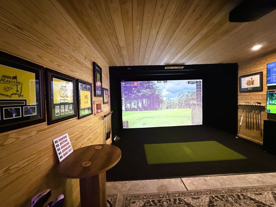 Indoor golf simulator setup in a room with wooden walls and a screen displaying a golf course.