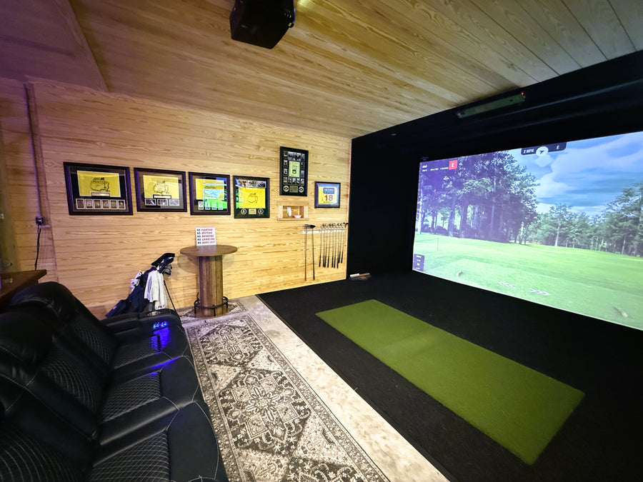 Indoor golf simulator setup with a screen displaying a golf course, surrounded by wooden walls and a rug.