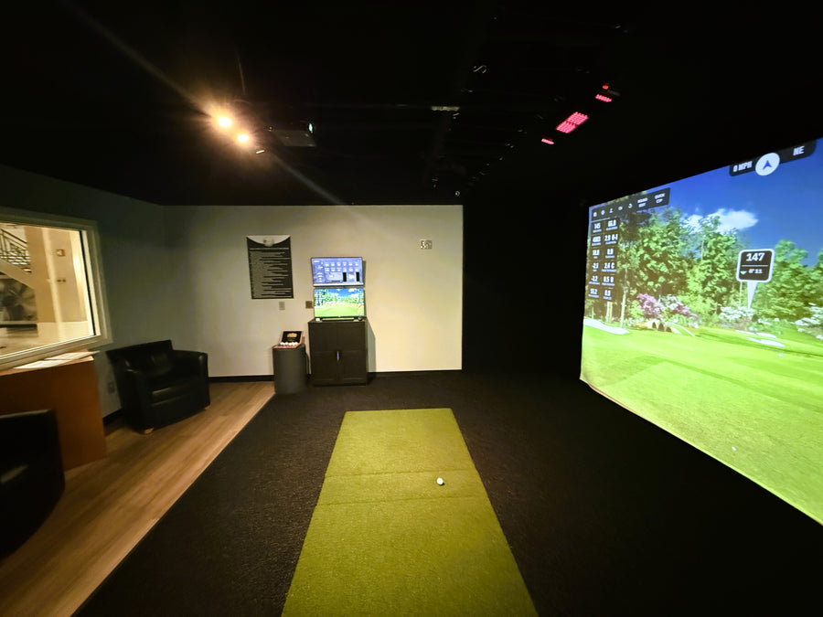 Golf simulator room with screen displaying a golf course and equipment on the floor.