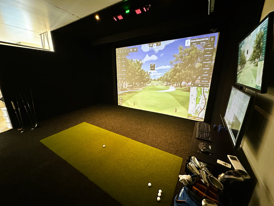 Golf simulator setup with screen displaying a golf course and various equipment on a mat.
