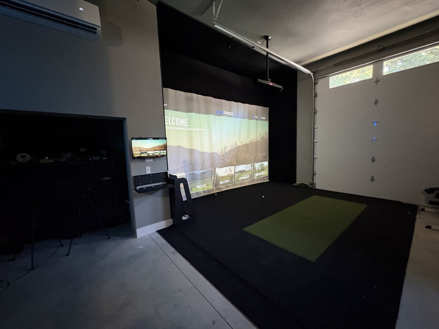 Garage with golf simulator and the screen displaying GSPro software. Green and black turf on the ground. 