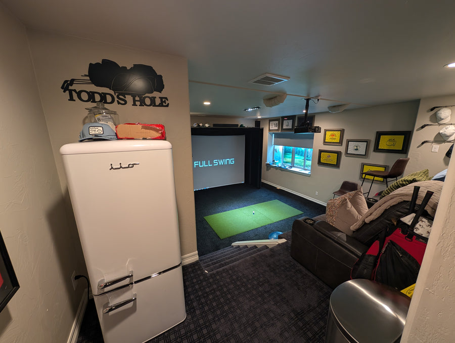 Indoor golf simulator setup in a room with a refrigerator labeled 'Todd's Hole'.