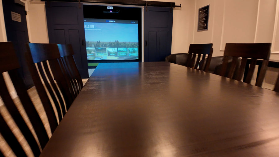 Long dark wooden table with chairs in a room with a screen and a golf simulator.